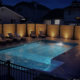 A backyard swimming pool at night is lit by warm lights along a wooden privacy fence, with lounge chairs and tables around the poolside. Nearby houses are visible, capturing the luxurious feel of custom infinity pools in Kelowna.