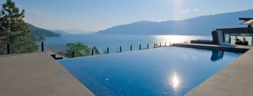 An infinity pool from the Luxury Pool Gallery overlooks a scenic lake and mountains under a clear blue sky, with sunlight shimmering on the water. A sleek glass fence lines the edge of this serene poolside retreat.