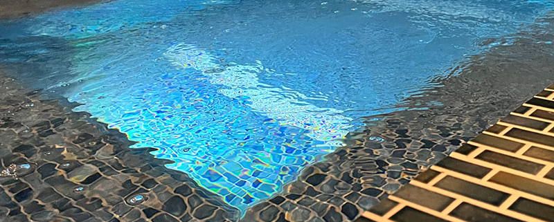 A close-up view of a tiled hot tub or small concrete pool at night, illuminated with blue underwater lights. The water surface reflects the lights, creating a shimmering effect against the dark, patterned tiles.