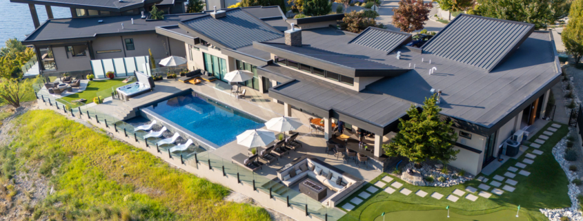 A modern lakeside house featuring an infinity pool design and installation, with lounge chairs, patio seating, umbrellas, a hot tub, and a small putting green—surrounded by landscaped greenery and overlooking the water.