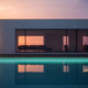 Modern house with large glass windows reflecting a pink and orange sunset sky, seen behind a tranquil Custom Concrete Pool. Minimalist interior furnishings are visible through the windows—perfect inspiration for pool construction near me.