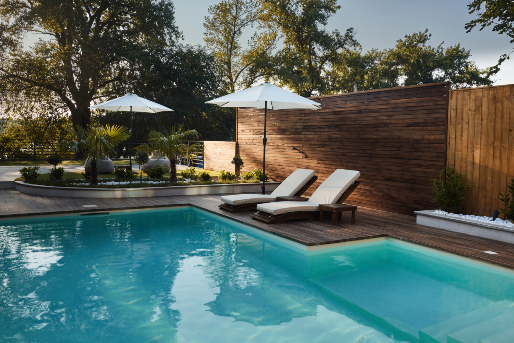 A modern outdoor swimming pool with clear water, expert Pool Installation, wooden decking, two cushioned lounge chairs under white umbrellas, and lush greenery in the background on a sunny day.