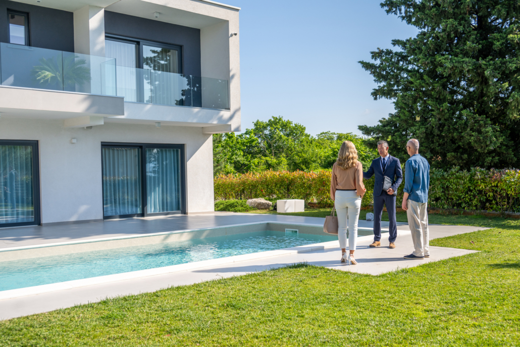 A man and a woman stand by a modern house’s pool, discussing Pool Installation options with a real estate agent in a suit on a sunny day, with trees and greenery surrounding the property.